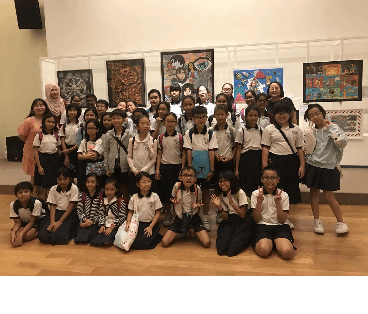 A large group of school children in white uniforms pose together in front of colorful artwork on gallery walls.